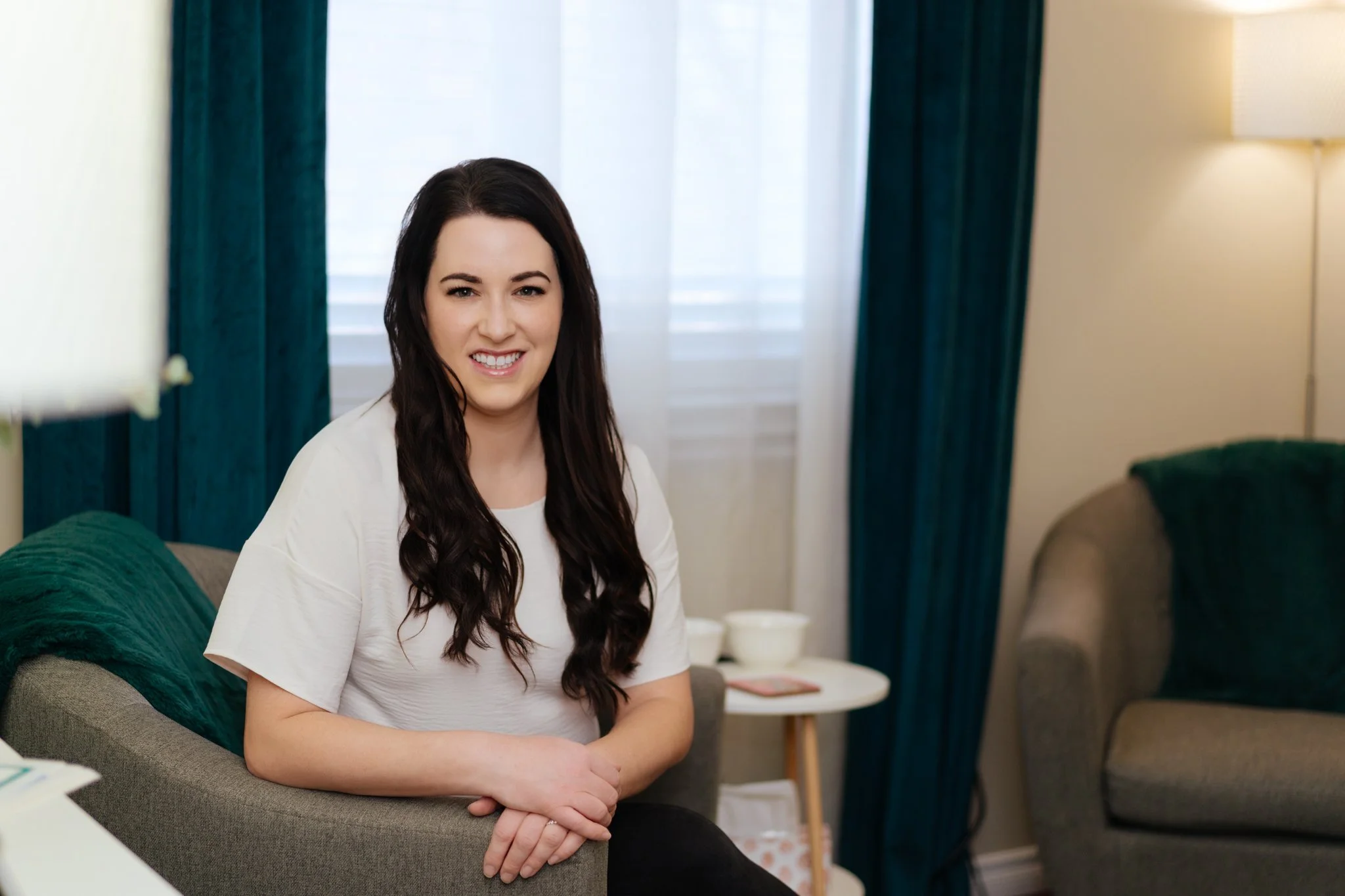 A woman with long dark hair smiling and sitting on a sofa in a well-lit living room with teal curtains and a side table.
