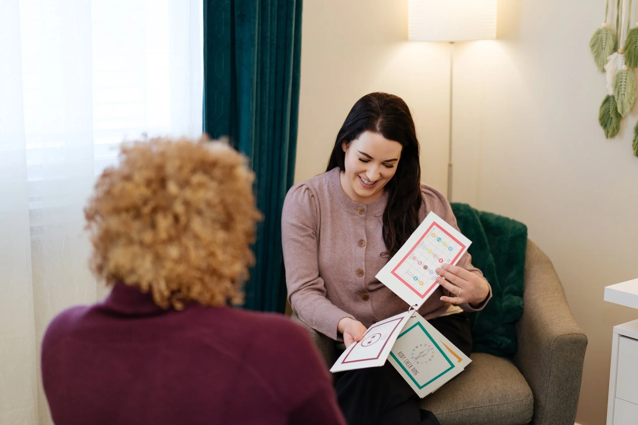 A woman with dark hair smiling while showing a colorful bingo card to an elderly woman with curly blonde hair in a cozy living room.