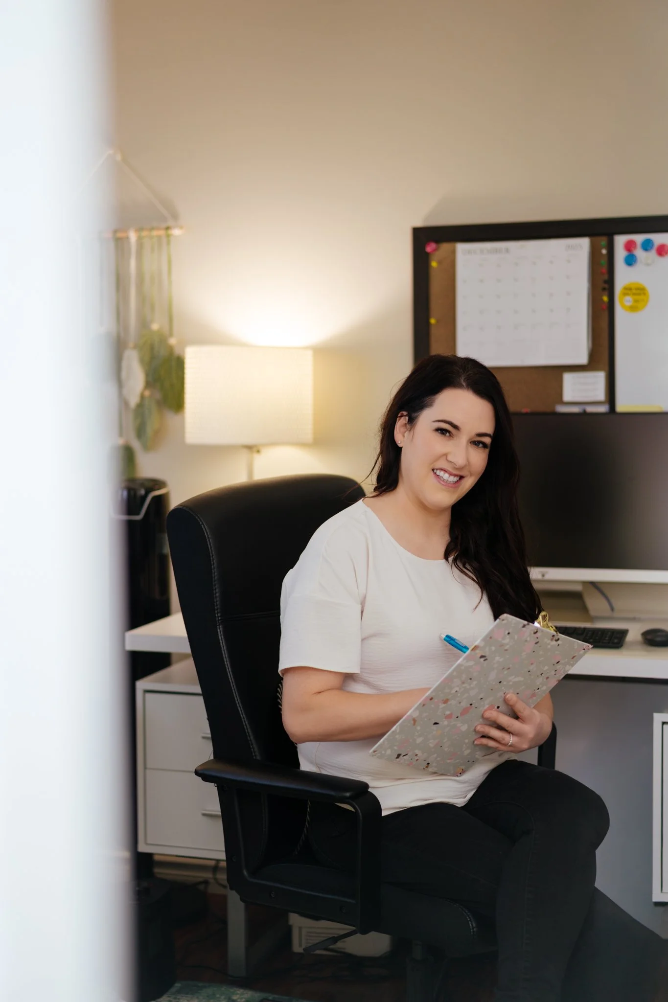A woman sitting in an office chair at a desk, smiling, holding a notebook and pen, with a computer monitor, a lamp, and a bulletin board in the background.