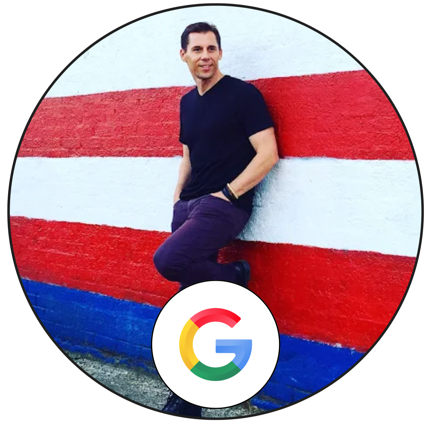 A man leaning against a colorful striped wall with red, white, and blue colors, smiling and dressed in a black t-shirt and dark pants.