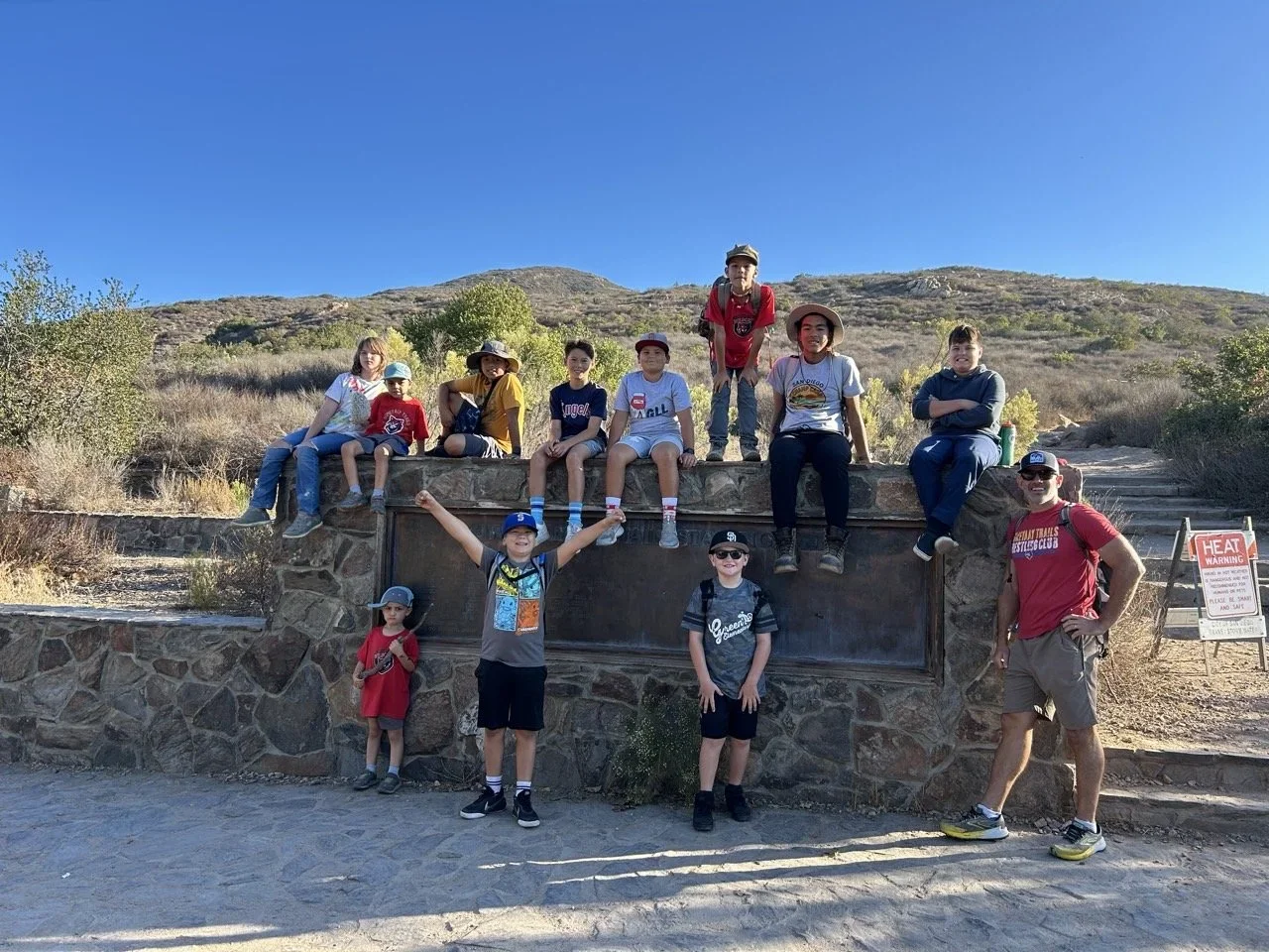 A group of children and two adults on a stone monument in a desert landscape with hills and sparse bushes, under a clear blue sky.