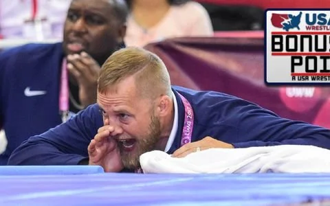 A wrestler lying on the mat, grabbing his face in distress, with a USA Wrestling bonus point sign in the background.