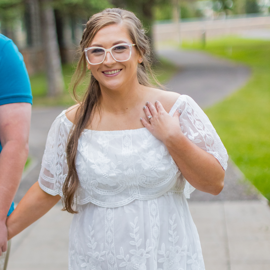 A smiling woman with glasses and long hair wearing a white embroidered dress, standing outside on a pathway with trees and grass in the background.