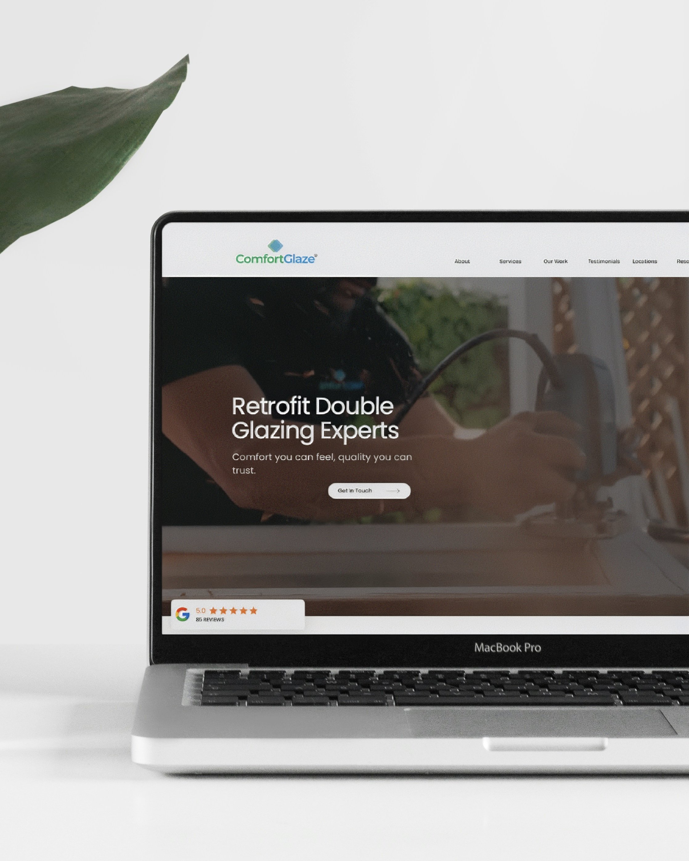 Laptop computer displaying a website for Comfort Glaze, a company specializing in retrofit double glazing, placed on a white surface with a green leaf in the background.