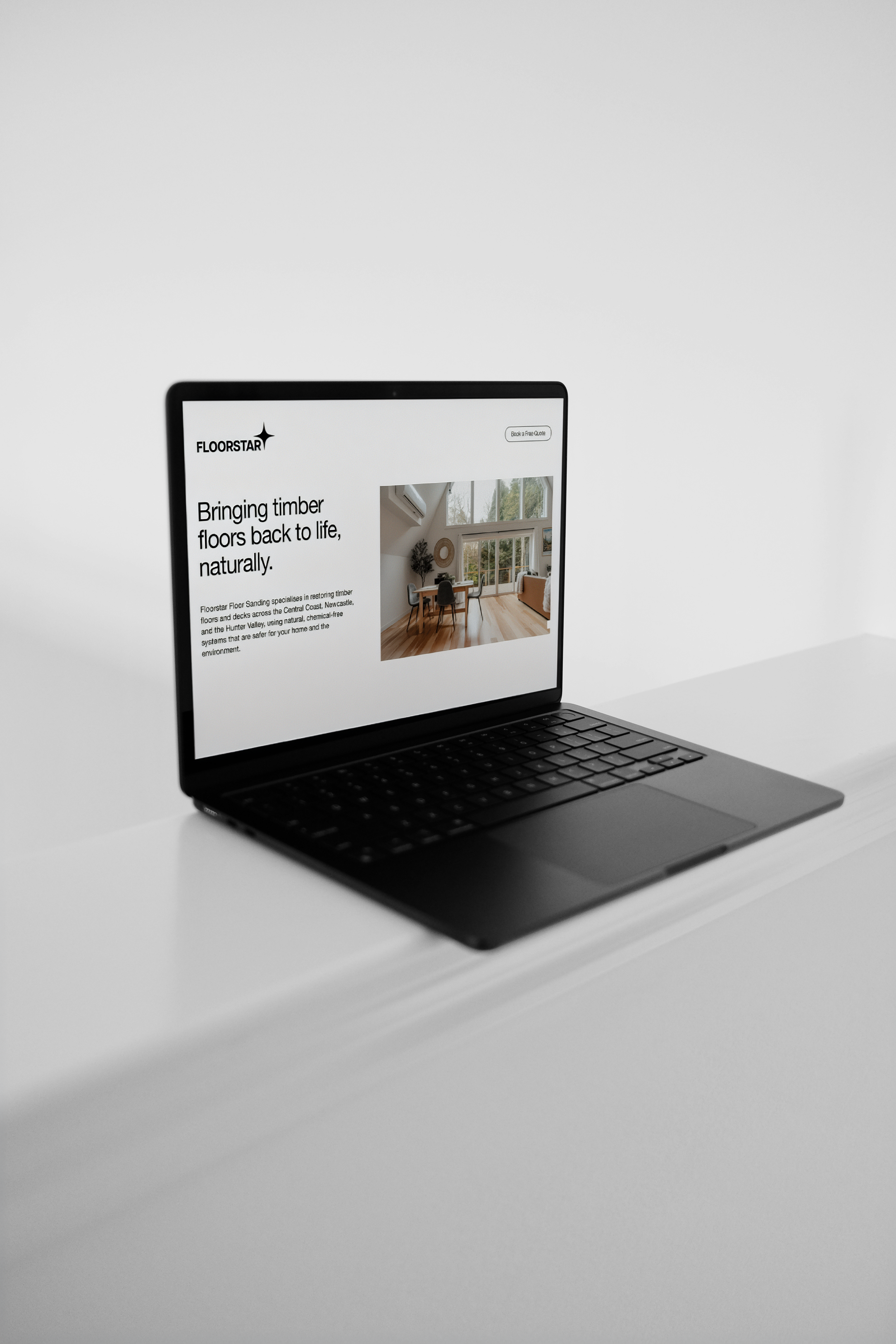 A black laptop on a white surface displaying a website about timber flooring, showing a living room with large windows and wooden floors.