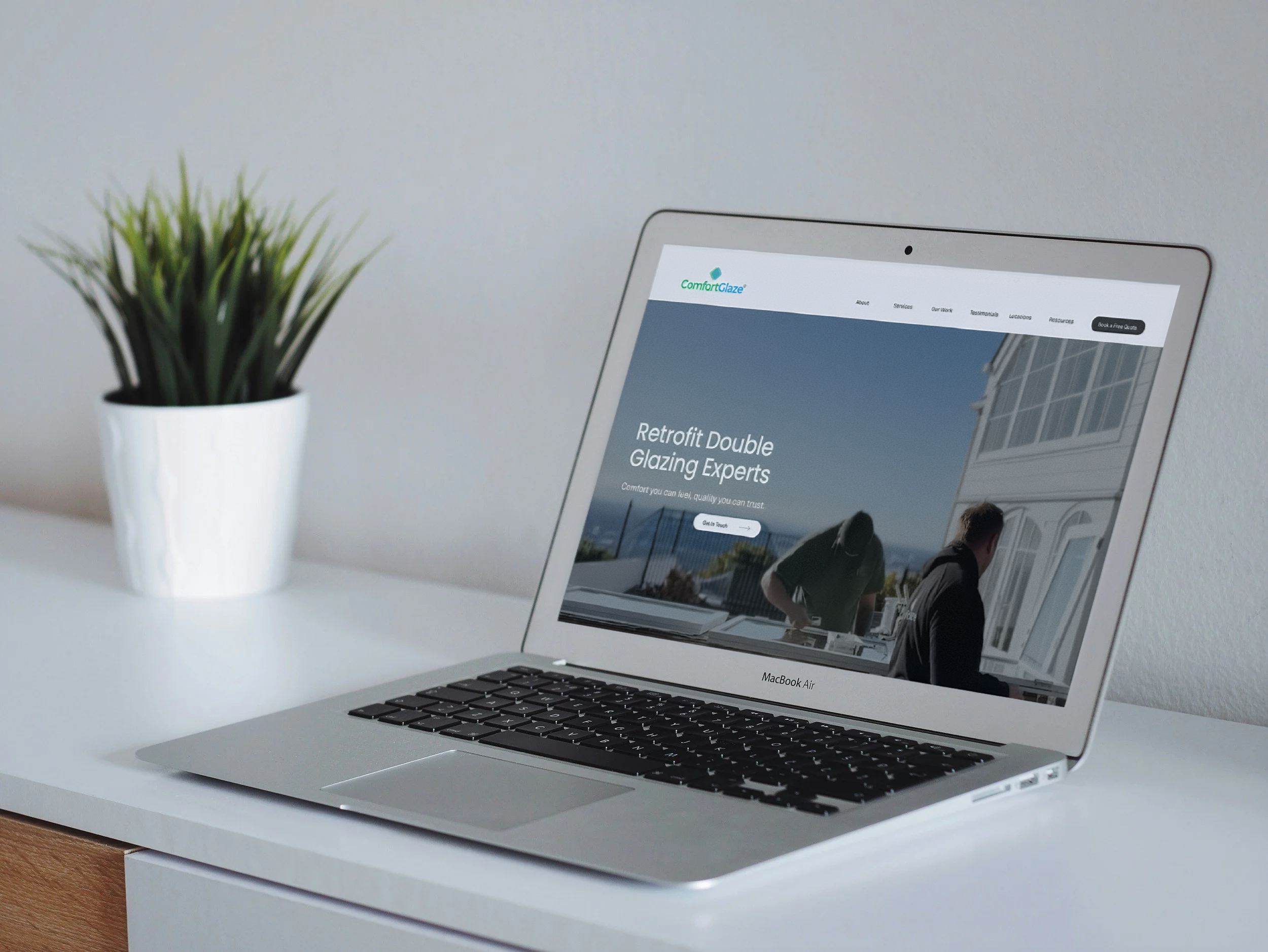 A silver laptop on a white desk displaying a website for ComfortGlaze with images of a house and people, next to a white pot with a green plant.