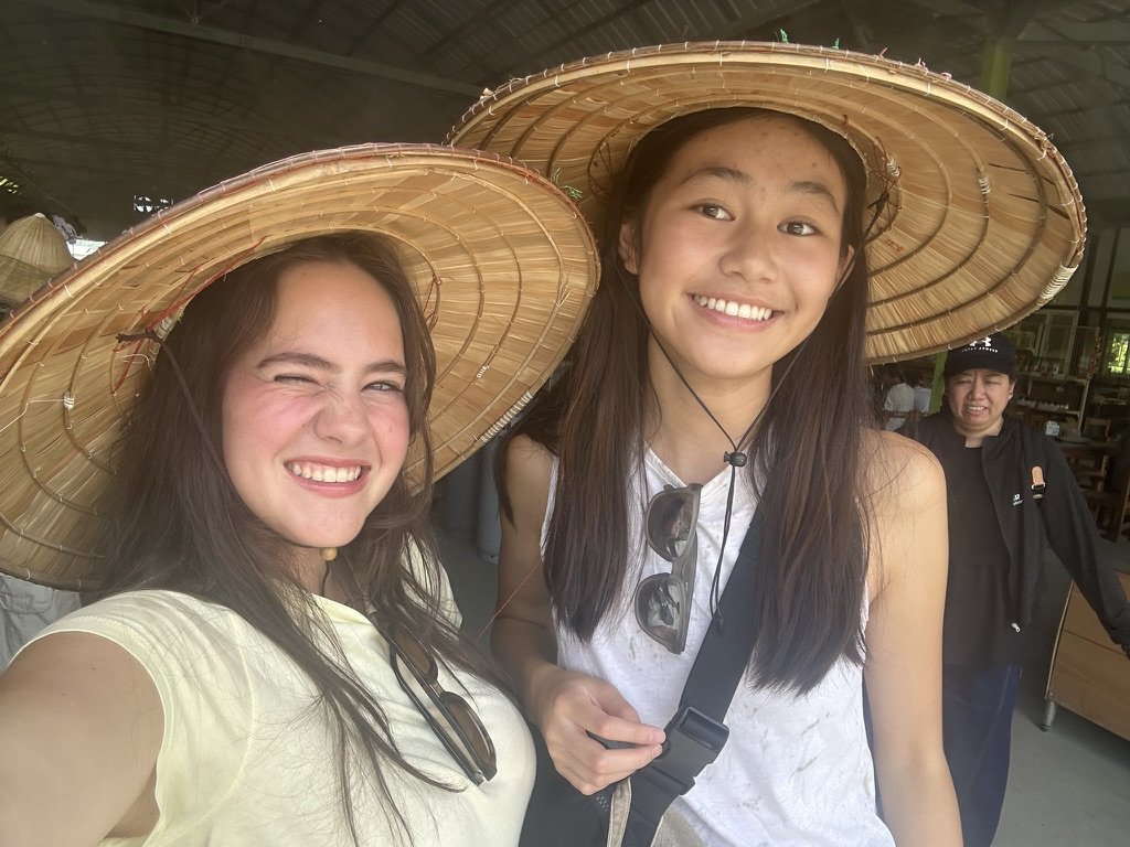 Two young women wearing wide-brimmed straw hats smiling for a selfie, with another woman in the background.