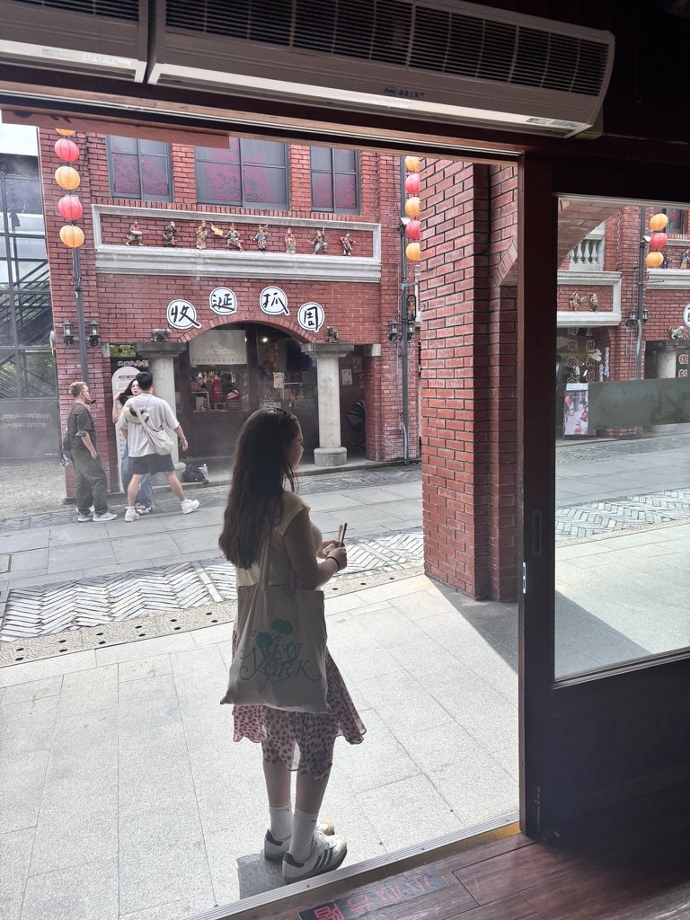 Young girl with long hair wearing a cardigan, skirt, socks, and sneakers stands inside a building looking out the glass door at a street scene with brick buildings, lanterns, and people walking.