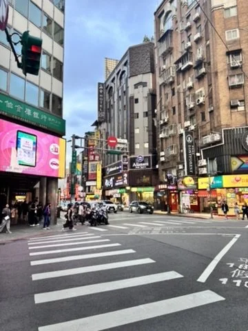 City street scene with pedestrians crossing at a crosswalk, tall buildings with various signs and storefronts, and a green traffic light in an urban setting.