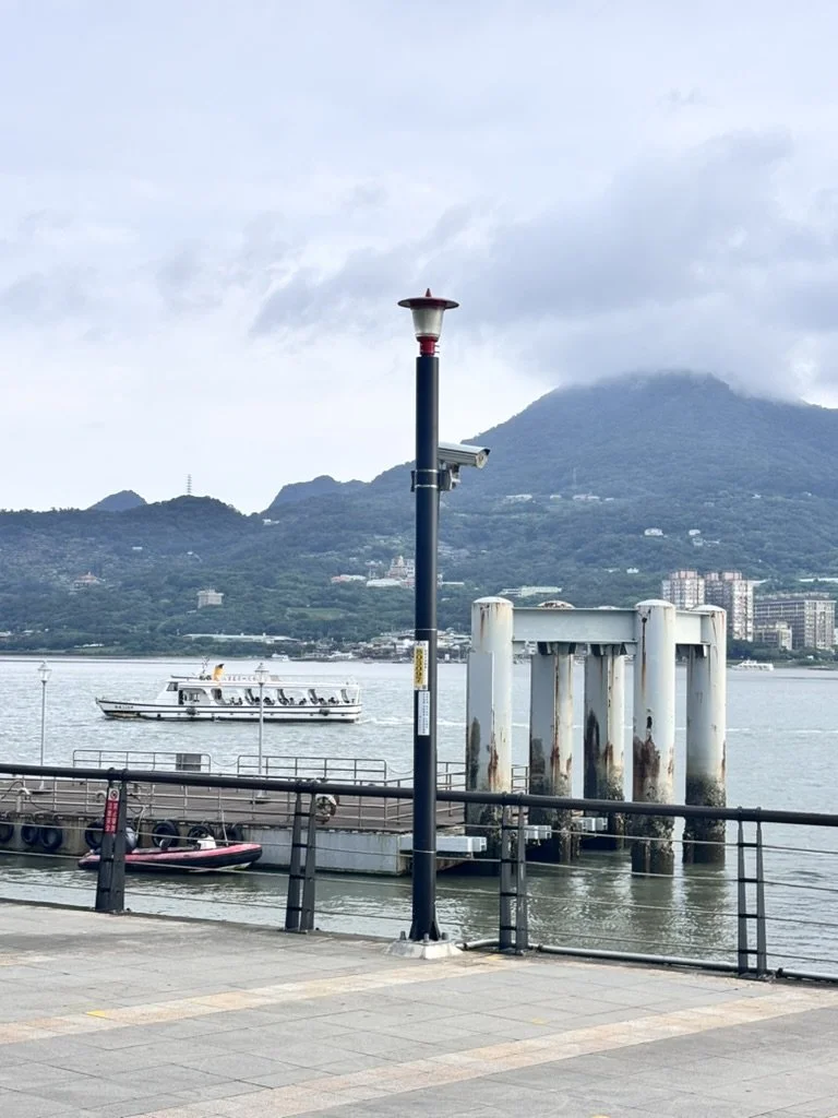 A waterfront scene with a lamp post and security camera, a boat in the water, and mountains in the background, under a cloudy sky.