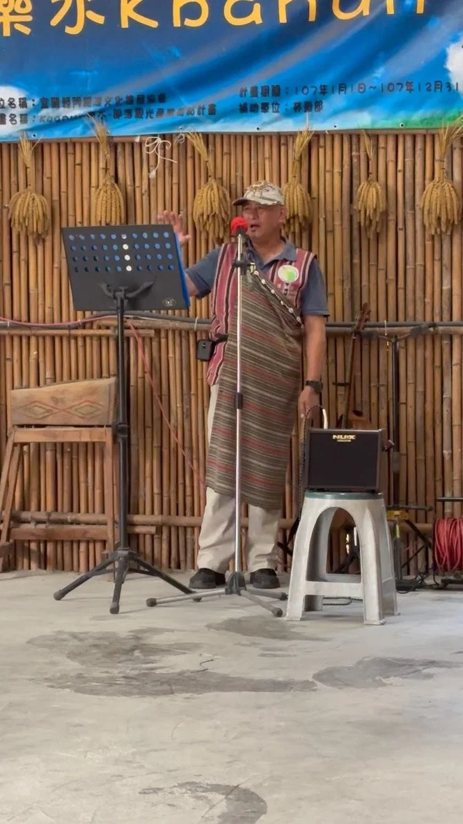 A person wearing a hat and traditional clothing standing in front of a bamboo backdrop, speaking into a microphone at an outdoor event.