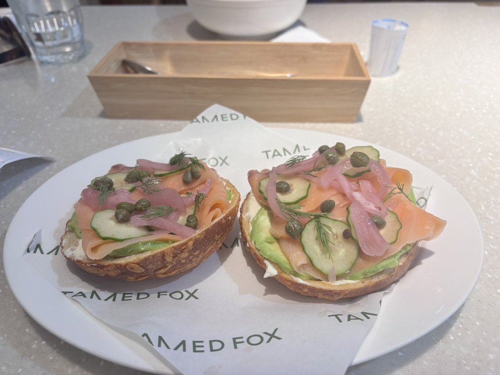 Two open-faced sandwiches with slices of cucumber, smoked salmon, pickled onions, fresh dill, and capers on toasted bread, served on a white plate with tarted FOX printed paper liner.