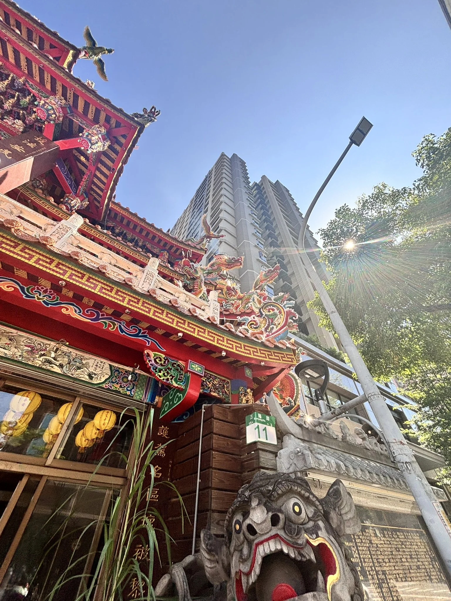 Colorful Chinese temple entrance with dragon statues, lion head sculpture, high-rise buildings in the background, streetlamp, and trees under a clear blue sky.