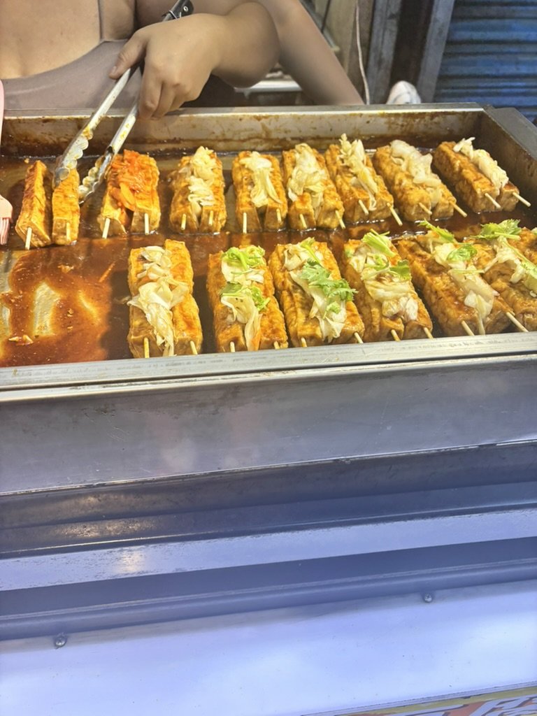 Street vendor preparing and selling grilled skewers of fried fish topped with lettuce, on a cooking grill with sauce.
