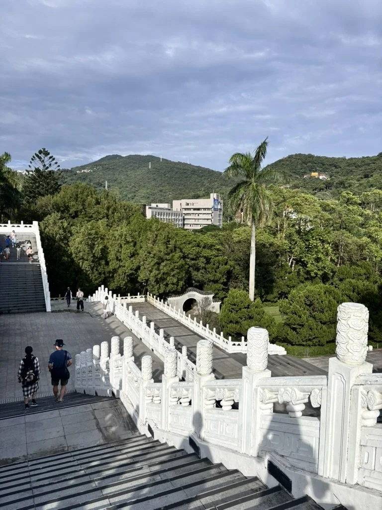 White marble stairs with ornate railing leading down to a park with trees, a tall palm tree, and hills in the background under a cloudy sky.