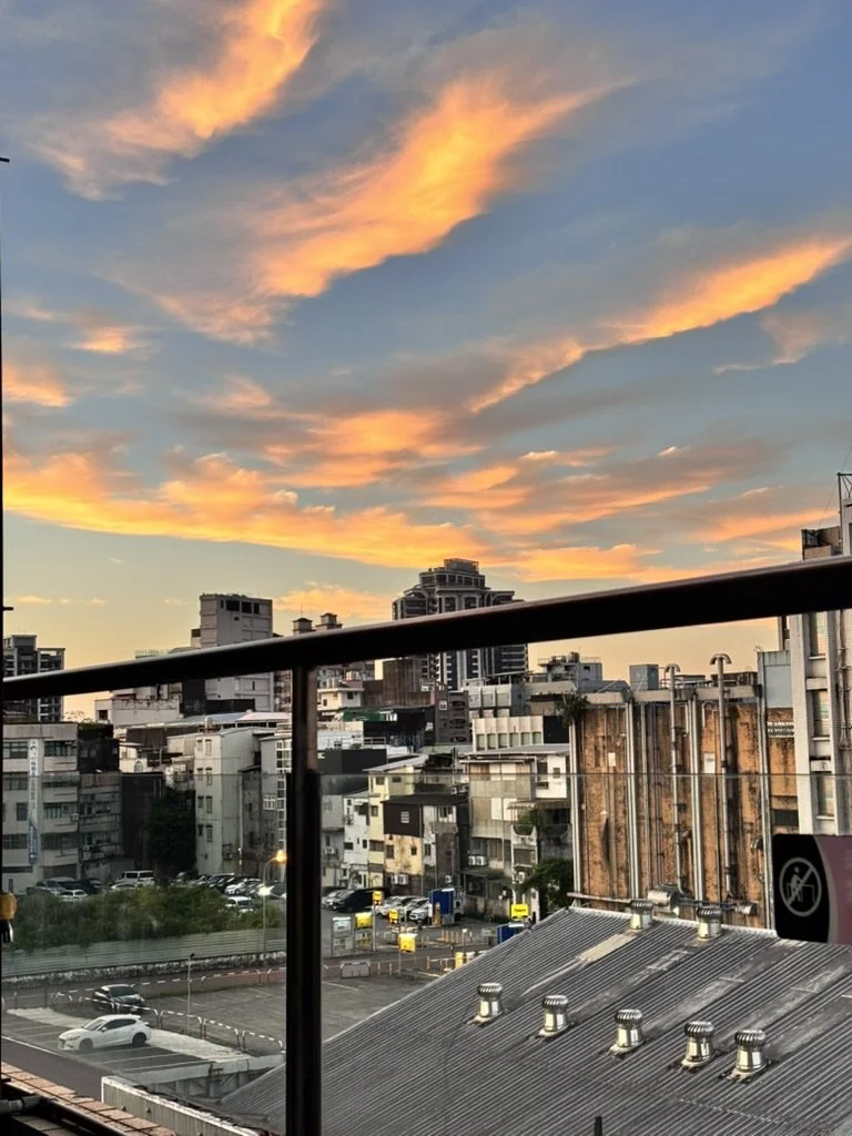 Cityscape during sunset with colorful orange and pink clouds in the sky over buildings, with a balcony railing in the foreground.