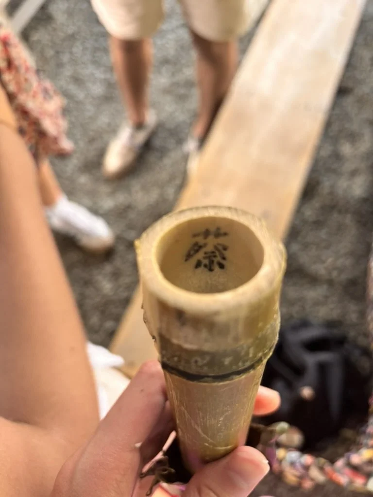 A person holding a bamboo flute with Chinese characters inside, with three people in the background standing on a gravel surface near a wooden bench.