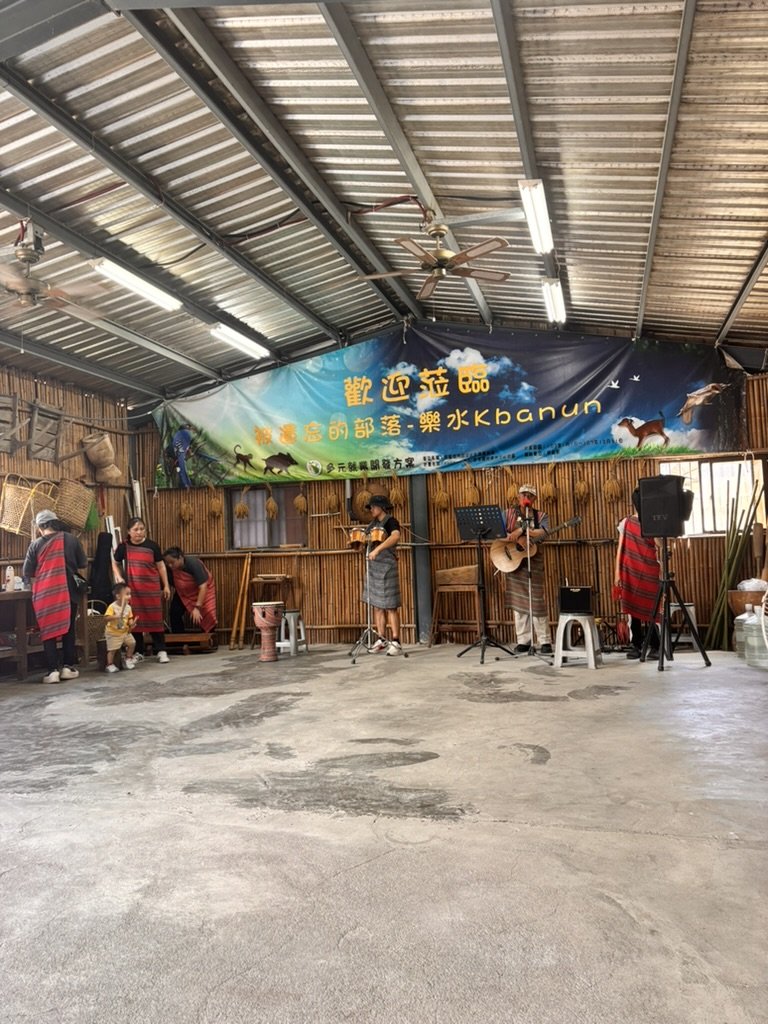 Group of people performing music with guitars in a rustic indoor setting, with a large colorful banner in the background featuring animals and Chinese characters.