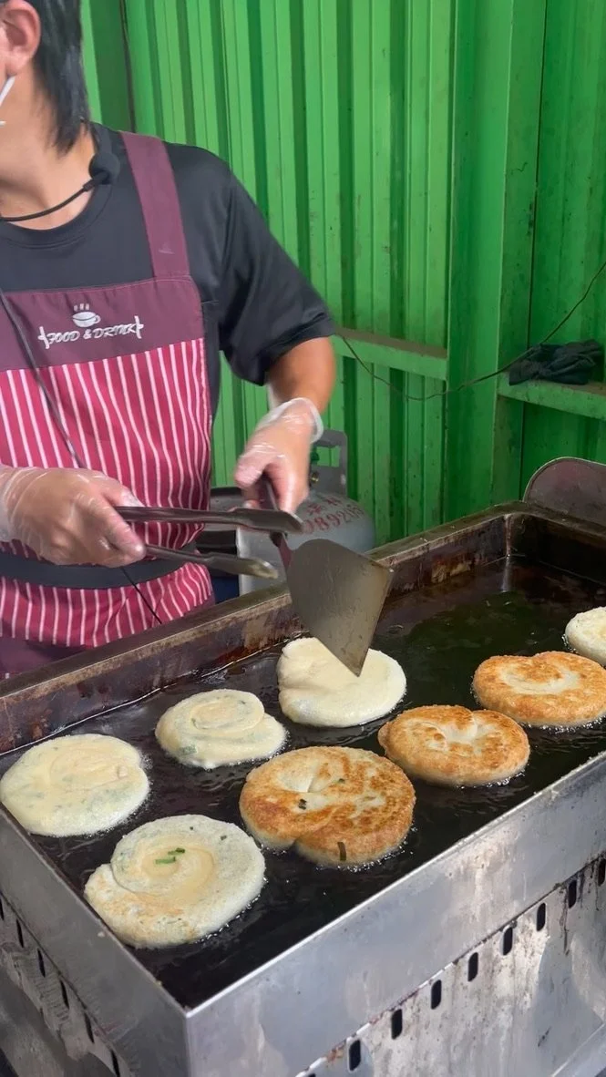 A person cooking scallion pancakes on a griddle, using tongs, in a green stall or kitchen. The pancakes are golden brown and some are still white.