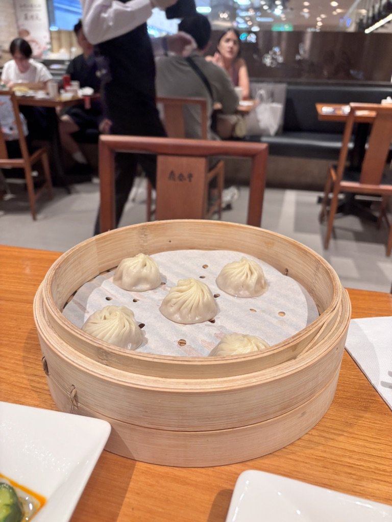 Six steamed dumplings in a round bamboo steamer on a wooden table in a restaurant, with other diners and waitstaff in the background.