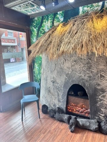 A realistic indoor fireplace with a thatched roof and stone exterior, surrounded by rocks, with an artificial fire burning inside. There is a blue chair nearby and a window showing a street view outside.