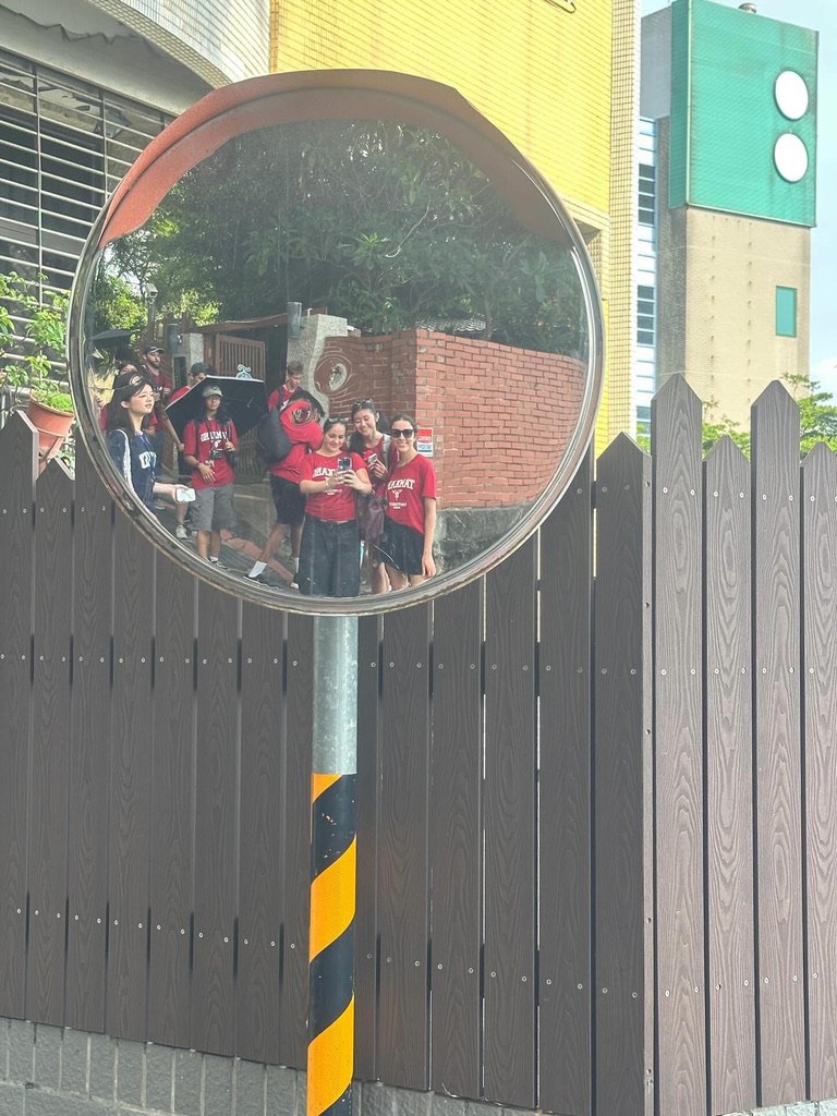 A group of people take a photo in a large circular mirror outside, reflecting a city scene with buildings and trees.