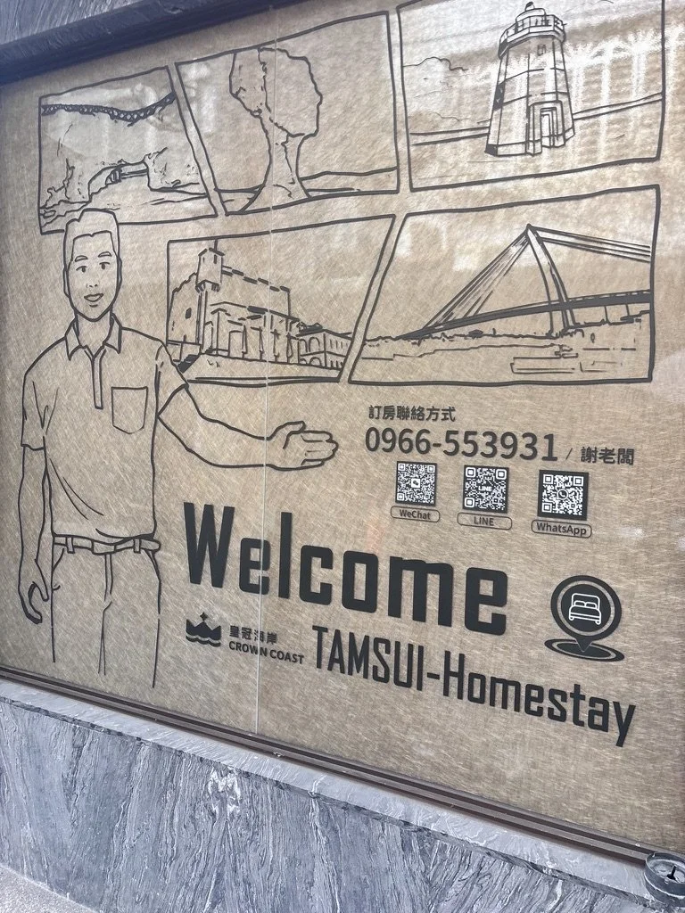 Signboard welcoming visitors to Tamsui Homestay with black and white line drawings of a bridge, a tree, a lighthouse, historic buildings, and a bridge over water. Includes contact phone number and QR codes for WeChat, LINE, and WhatsApp.