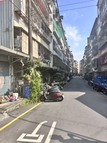 Narrow city street with parked scooters and cars, surrounded by multi-story residential buildings under a blue partly cloudy sky.