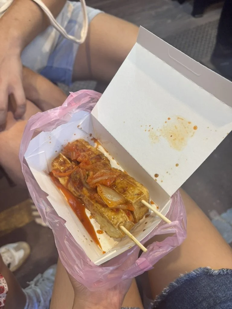 Person holding a takeout container with skewered Chinese food, likely egg roll stuffed with vegetables in a sauce with onion and peppers, in a pink plastic bag.