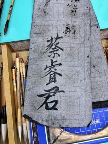 A piece of fabric with Japanese calligraphy, surrounded by pens, brushes, and writing tools on a cluttered desk.