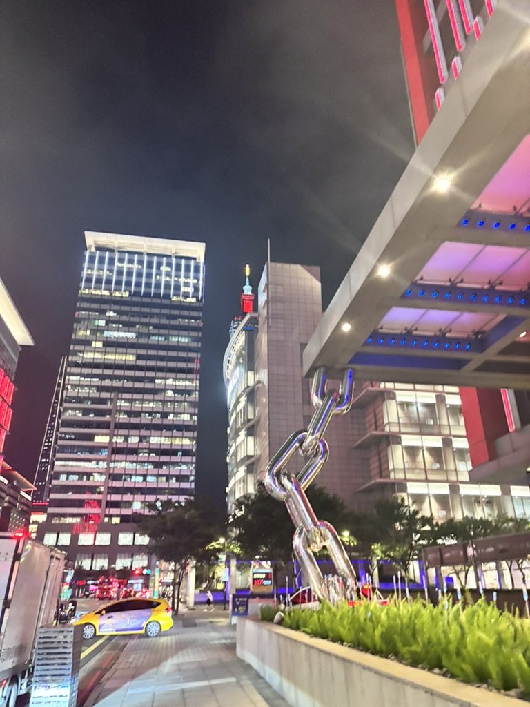 Nighttime city scene with illuminated skyscrapers, a large outdoor chain sculpture in the foreground, and colorful neon lights.