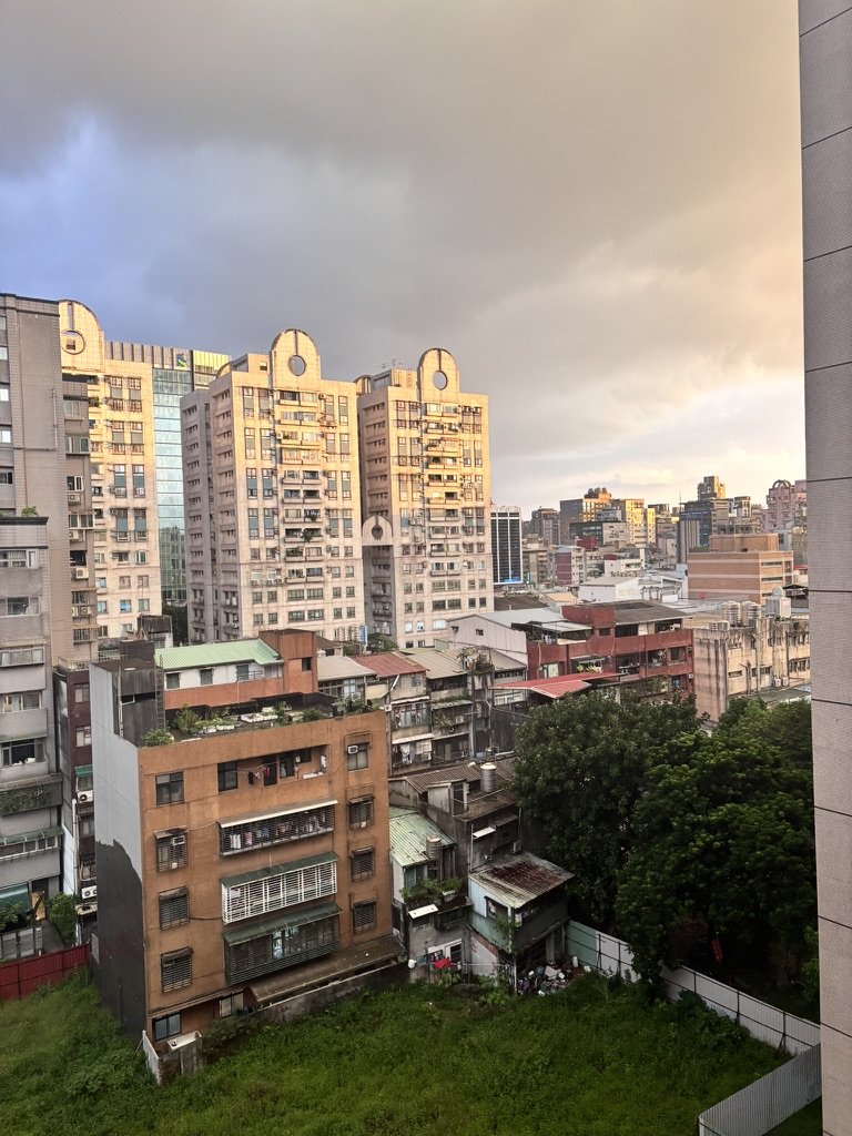 Cityscape with tall residential buildings and lower structures under a cloudy sky at sunset.