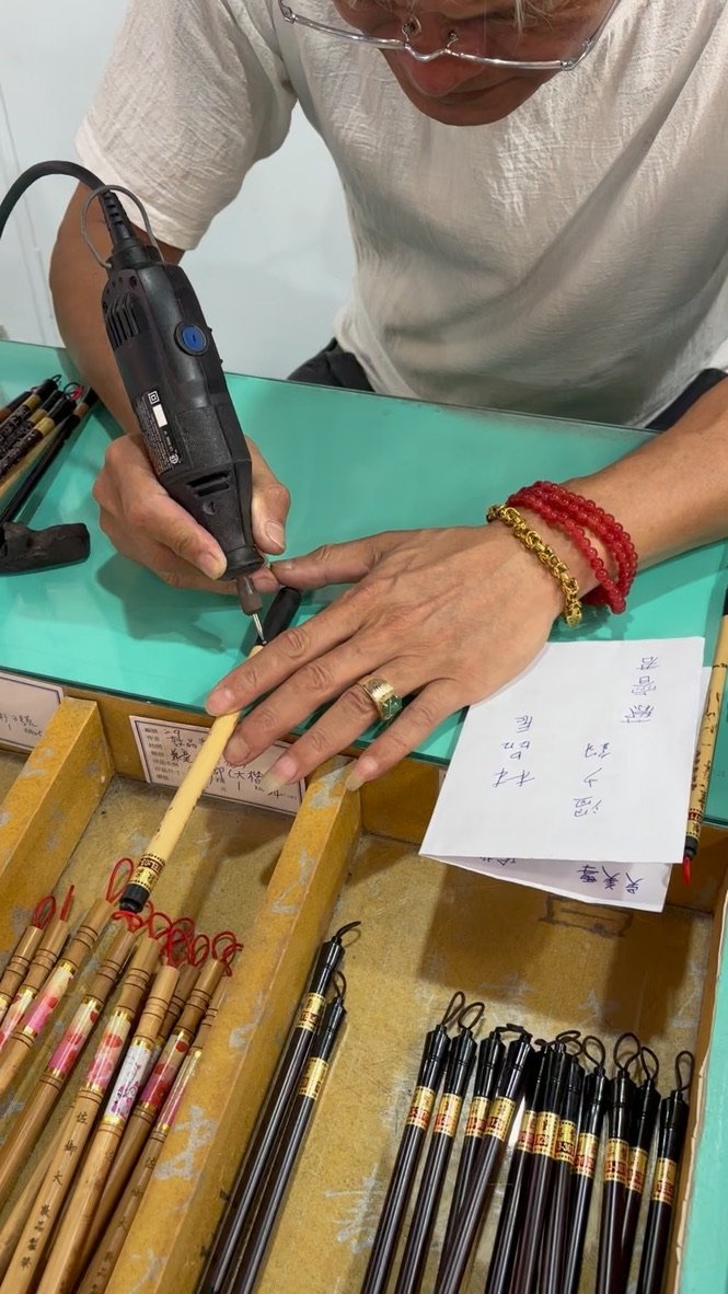 Person using a heated tool on a calligraphy brush at a desk with various other calligraphy brushes and papers with Chinese characters.