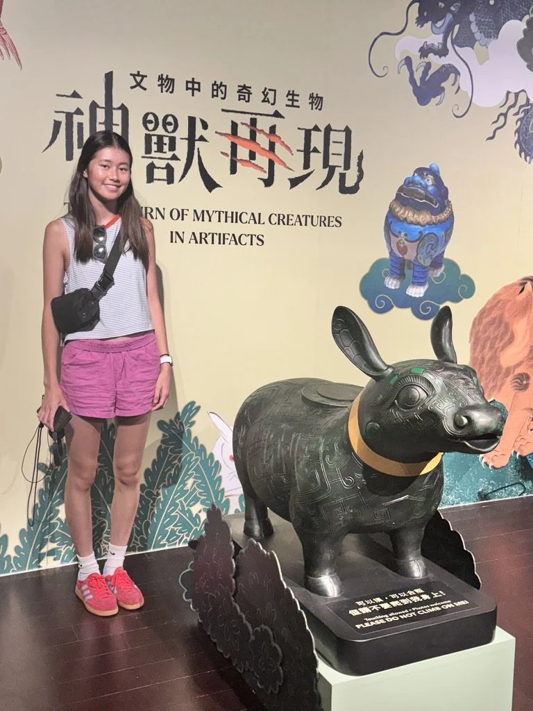 A woman with a black crossbody bag, wearing a sleeveless top, pink shorts, and pink shoes, standing next to a large black animal sculpture with rabbit ears at an exhibit about mythical creatures in artifacts.