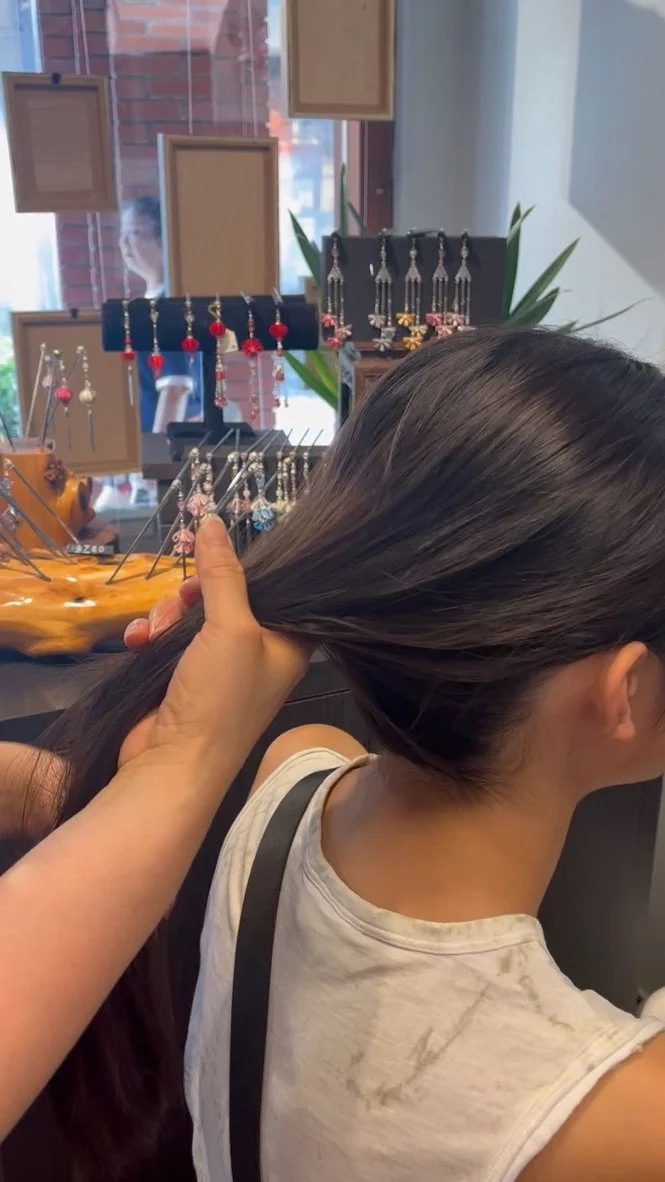 A woman getting her hair combed at a jewelry store, with display earrings and framed pictures in the background.