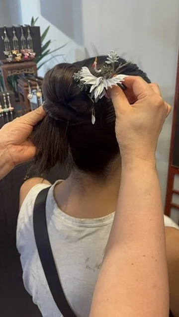 A person with dark hair is having a decorative hair accessory placed in an updo. The accessory looks like a floral headpiece with white and green elements.