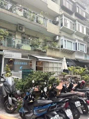 A row of motorcycles and scooters parked outside a building with a sign that reads 'SEL SHUI', with balconies and greenery above.