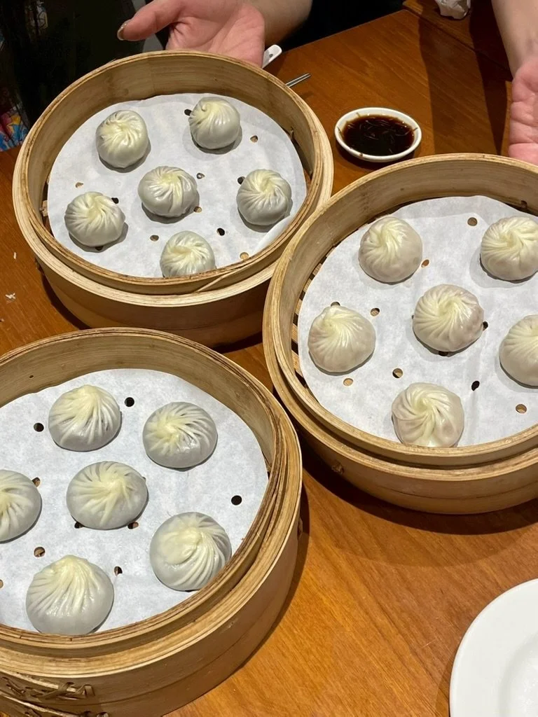 Several bamboo steamers containing steamed dumplings on a wooden table, with a small dish of soy sauce nearby.