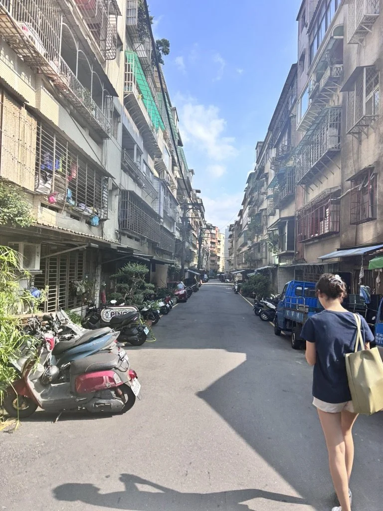 A street scene with parked scooters and motorcycles on the left, residential apartment buildings on both sides, and a young woman walking away from the camera on the right. The sky is blue with some clouds.