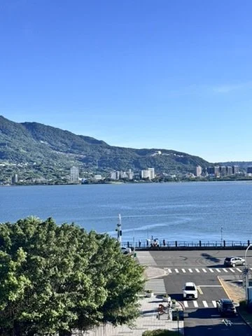 View of a lake with mountains in the background, a cityscape along the shoreline, a parking lot, and green trees in the foreground.
