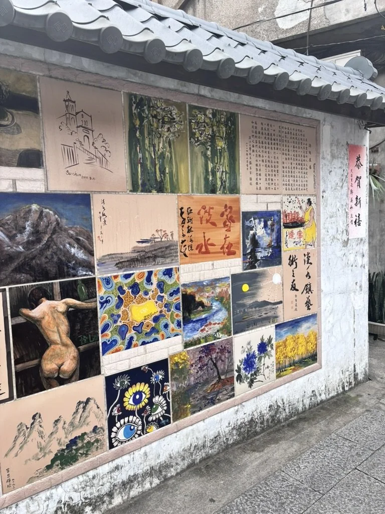 A mural wall with various paintings, drawings, and Chinese calligraphy, including landscapes, flowers, and a portrait of a woman in traditional attire.