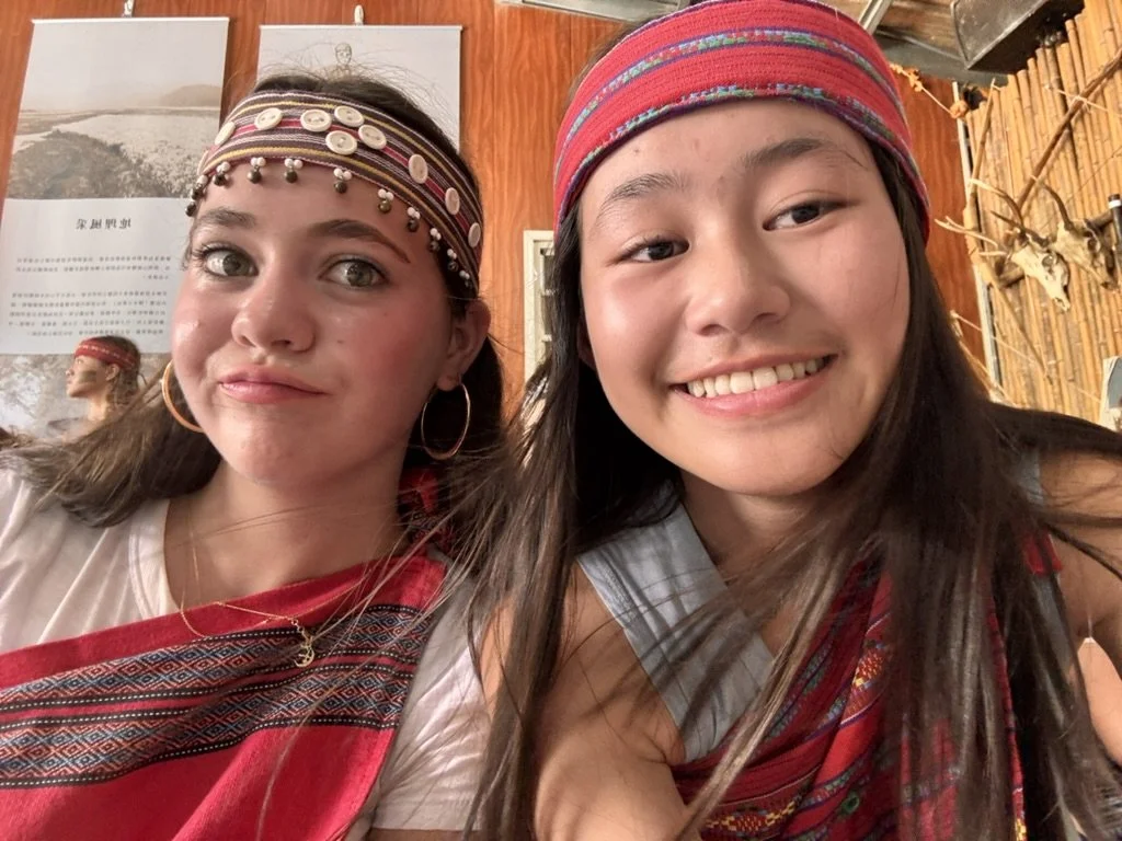 Two young women smiling and wearing colorful, traditional cultural accessories and headscarves inside a wooden-walled room decorated with animal skulls and photographs.