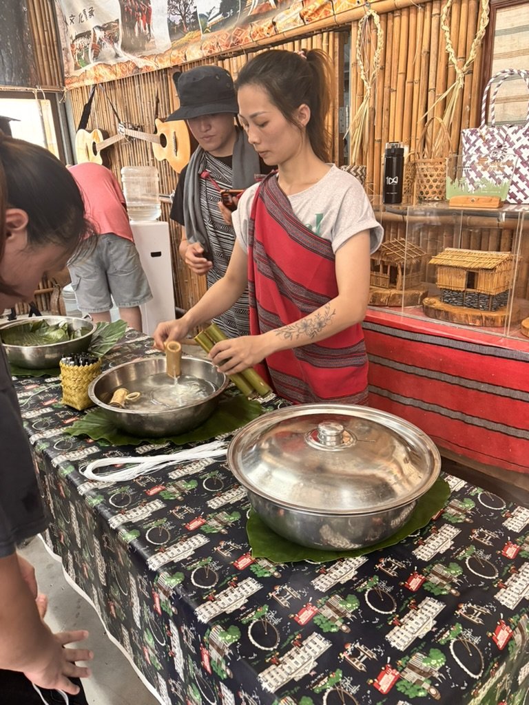 A young woman with a tattoo on her arm, wearing a gray T-shirt and a red and black striped shawl, prepares traditional food with bamboo tubes at a market stall. Two other women are nearby, one in a black hat and gray scarf, and another partially visi