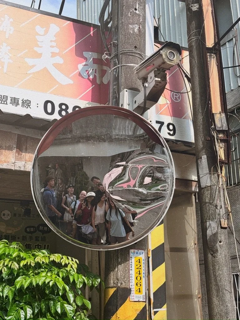 A group of people reflected in a convex traffic mirror mounted on a pole at an urban street corner. The mirror is in front of a utility pole with a surveillance camera mounted above it and a sign with yellow and black stripes at the pole's base. Buil