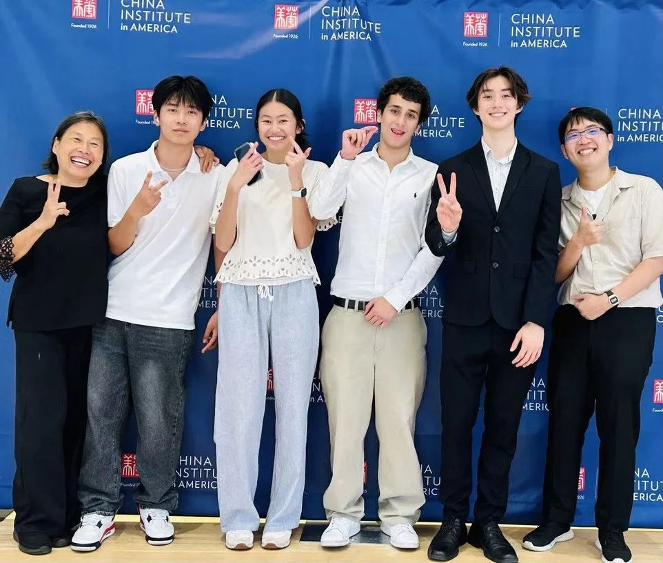 Group of six young people and one adult smiling and making peace or victory signs in front of a blue backdrop with China Institute in America logo.