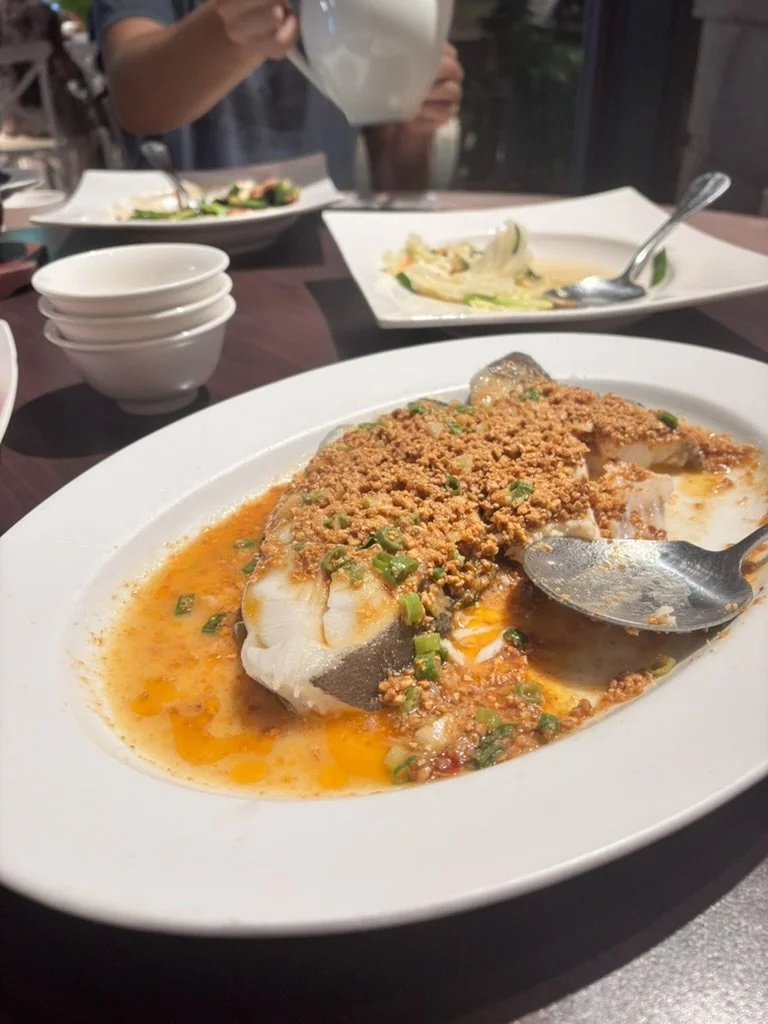 A white oval plate with fish topped with crushed peanuts and green onions, served in a spicy orange sauce. In the background, there are broken rice and vegetables on white dishes, and a person pouring tea.