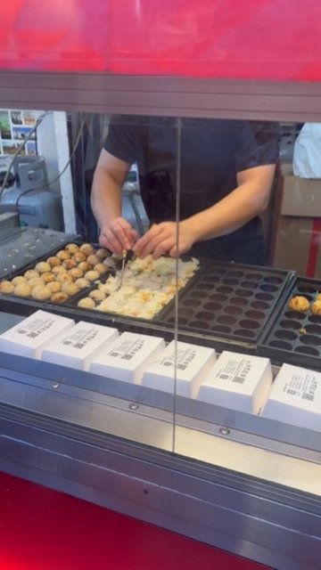 A person making takoyaki, Japanese octopus balls, at a food stall with a takoyaki grill and boxes of ingredients.