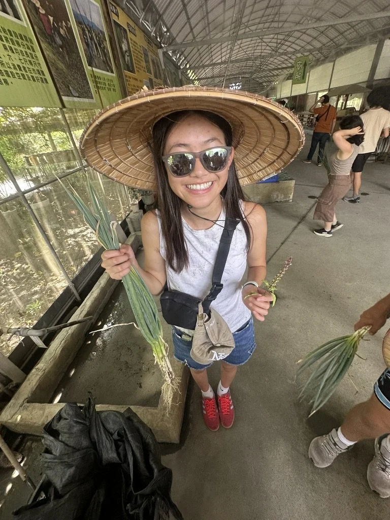 A girl in sunglasses wearing a large traditional straw hat, holding green plants, standing inside a greenhouse or agricultural facility, smiling at the camera.