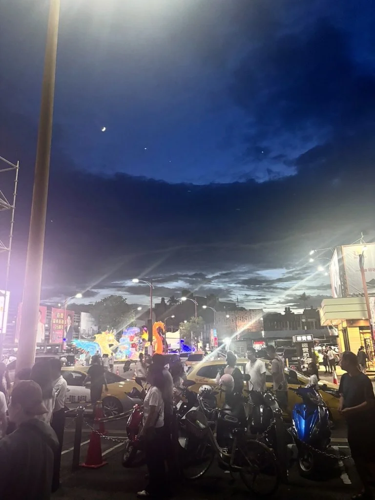 A lively city street scene at dusk with streetlights, colorful illuminated sculptures, taxis, motorcycles, and pedestrians under a darkening sky with a visible moon.
