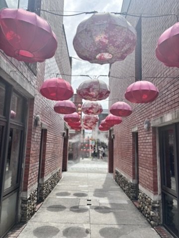 Narrow alleyway lined with brick buildings, decorated with pink and white lanterns hanging overhead.